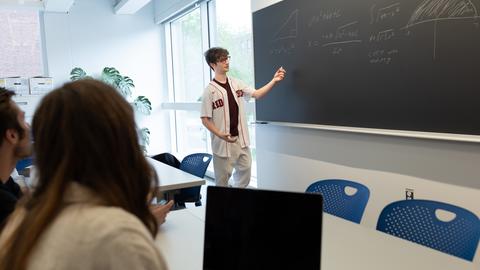 A student explains math at a chalkboard while another takes notes on a laptop.