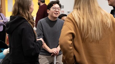 Four people standing in a circle, having a conversation. One person faces the camera, leaning on crutches with both hands and smiling while conversing.