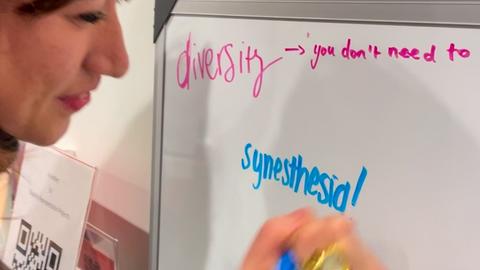 A woman writes  "synesthesia!" on a whiteboard. Also written is "Diversity: you don't just need to..."