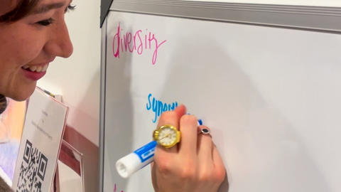 A woman is writing the word "synesthesia" on a whiteboard in blue. "Diversity" is written in pink in the corner of the whiteboard.