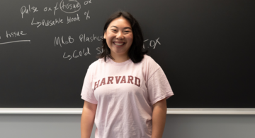 Person wearing a Harvard shirt smiling in front of black chalkboard