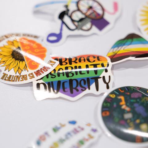 Stickers on a table, the one in focus reading "Embrace Disability Diversity" against a multi-colored heart.