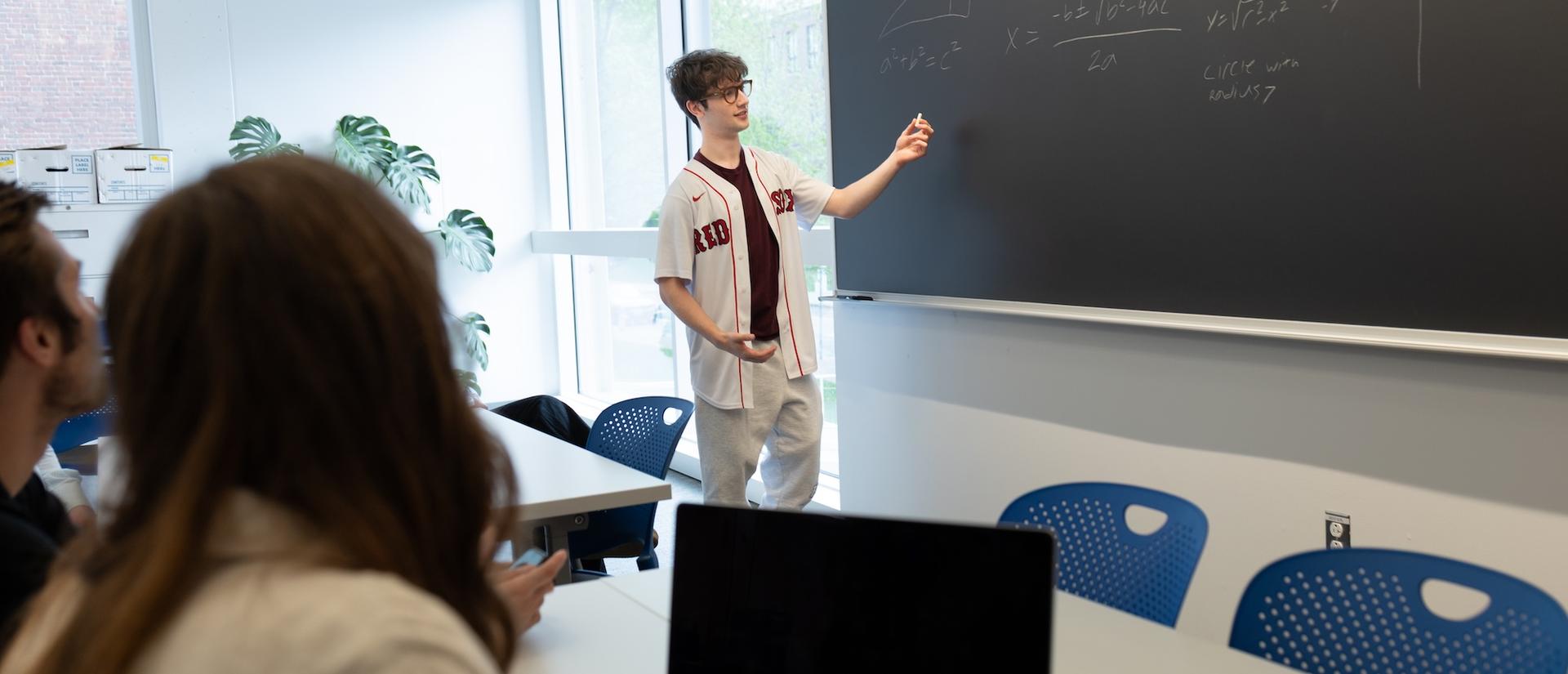 A student explains math at a chalkboard while another takes notes on a laptop.