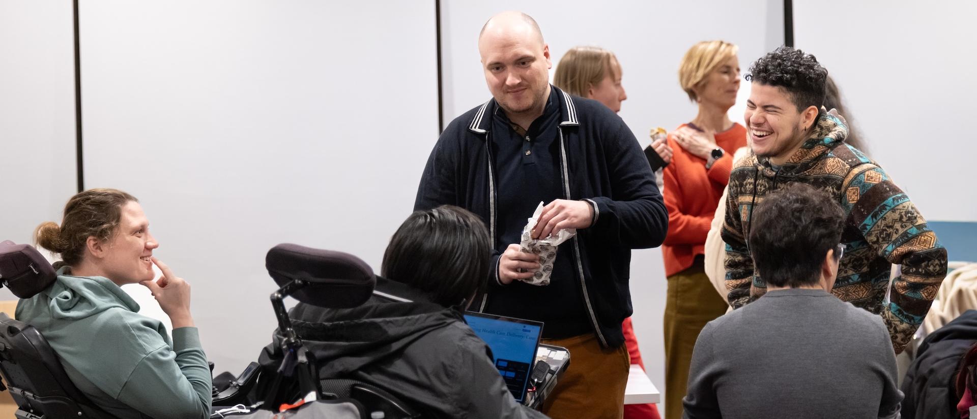 Several people laughing and having a conversation in a classroom. A few are in wheel chairs, others are standing.