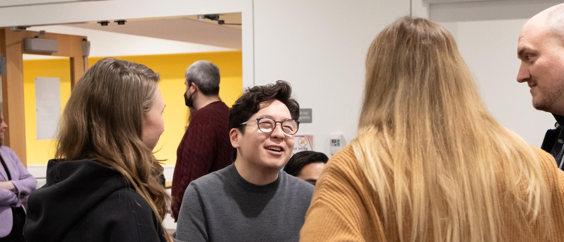 Four people standing in a circle, having a conversation. One person faces the camera, leaning on crutches with both hands and smiling while conversing.