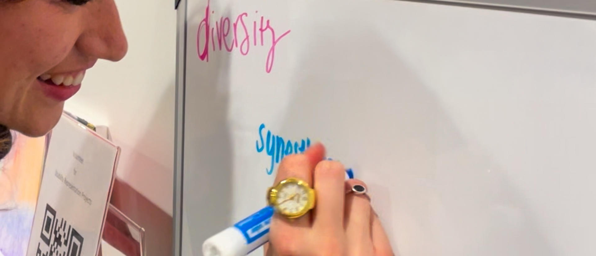 A woman is writing the word "synesthesia" on a whiteboard in blue. "Diversity" is written in pink in the corner of the whiteboard.